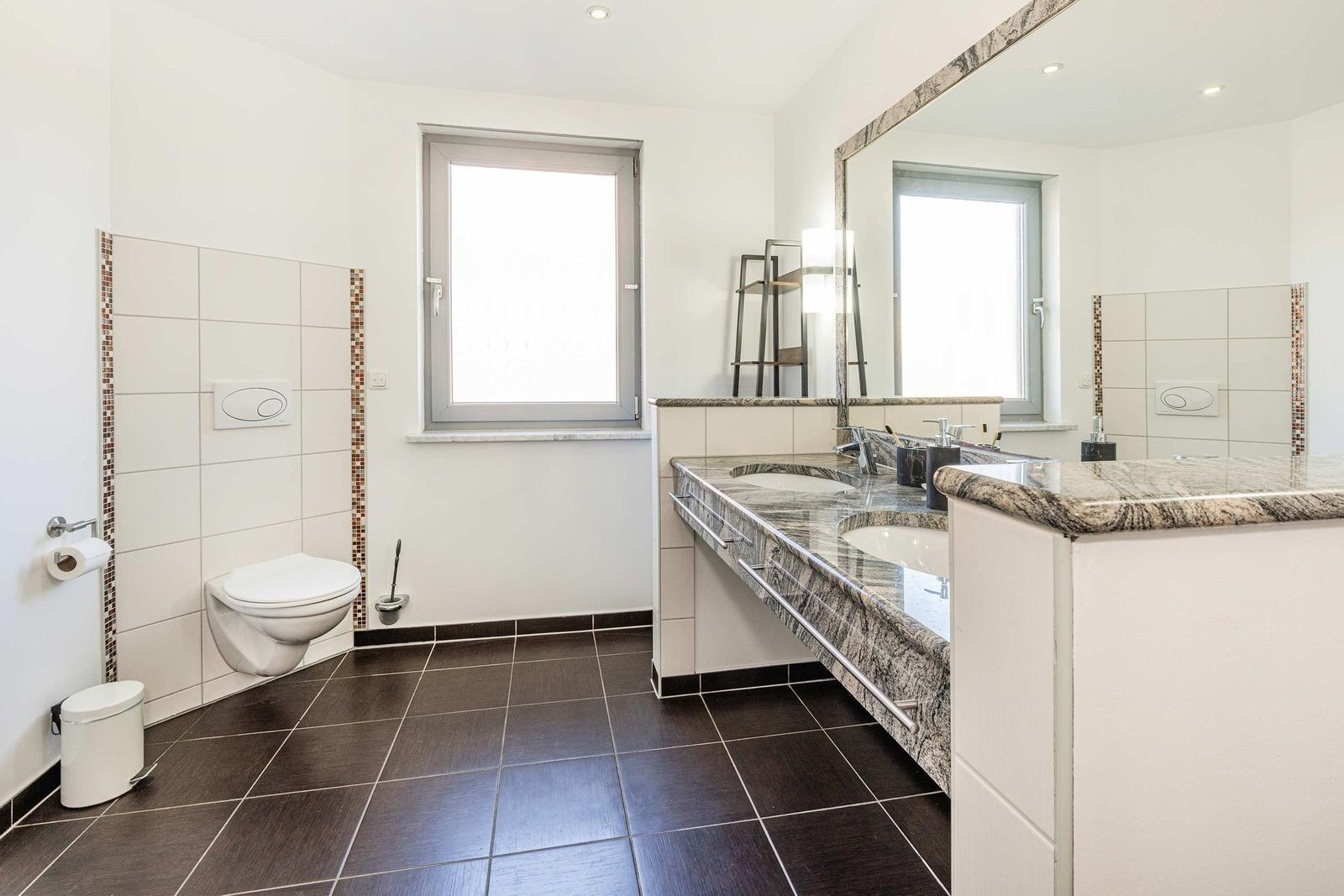 Badezimmer mit Doppelwaschbecken, Spiegel und Toilette. Bodenfliesen und Wandfliesen.
