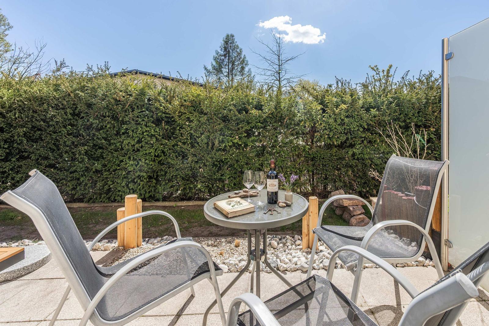 Terrasse mit Tisch und Stühlen, Weinflasche und Blumen vor grüner Hecke.