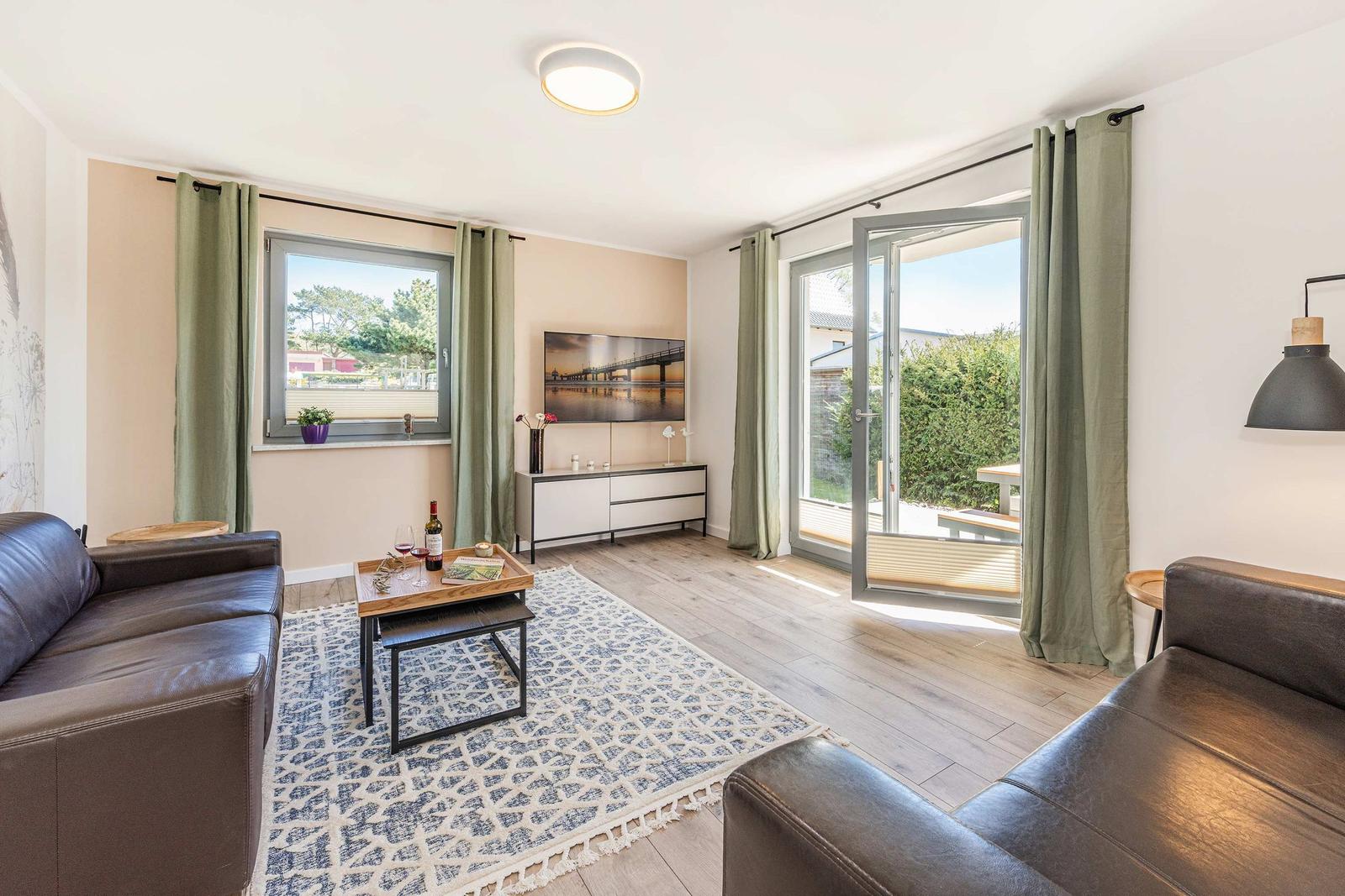Wohnzimmer mit Sofa, Fernseher und Terrasse. Grüne Vorhänge und Holzboden.