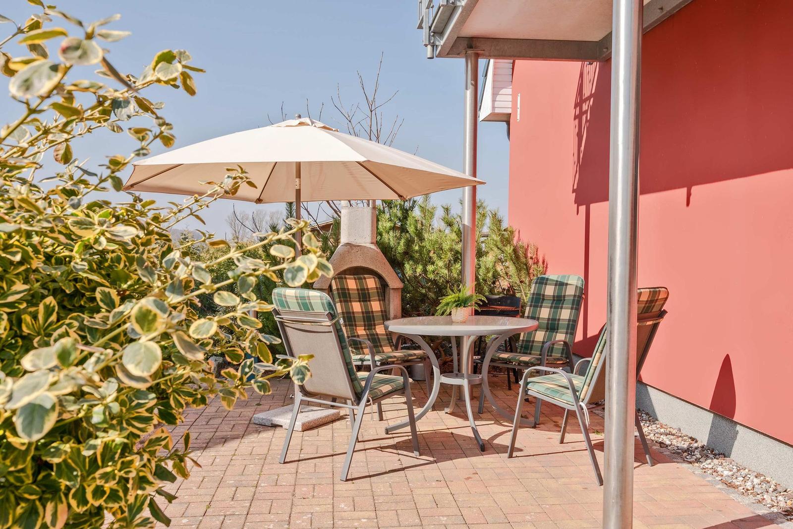 Terrasse mit Tisch, Stühlen, Sonnenschirm und Grillofen. Rotes Gebäude und Pflanzen im Hintergrund.