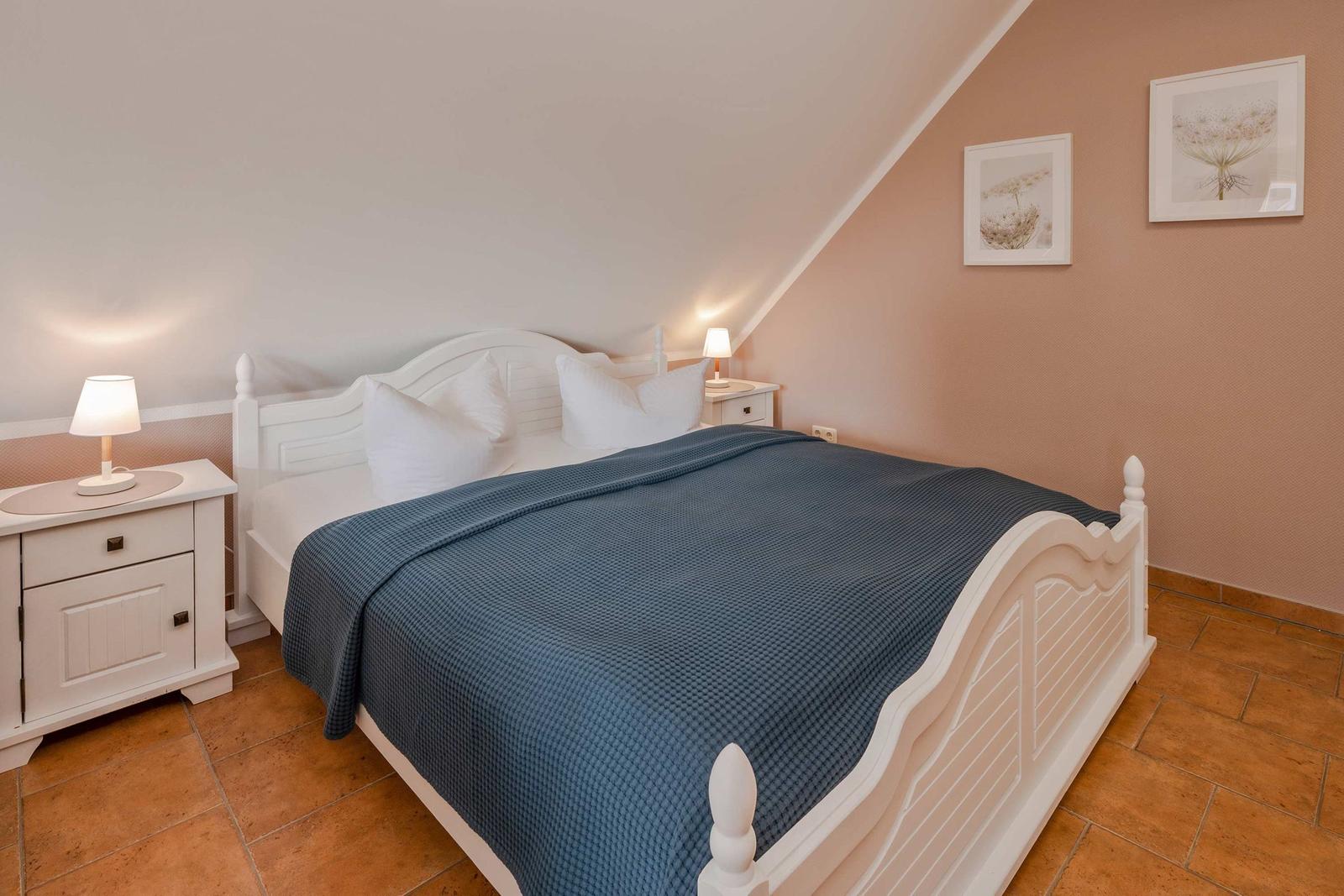 Schlafzimmer mit weißem Bett, blauer Bettdecke, zwei Nachttischen und zwei Bildern an der Wand.
