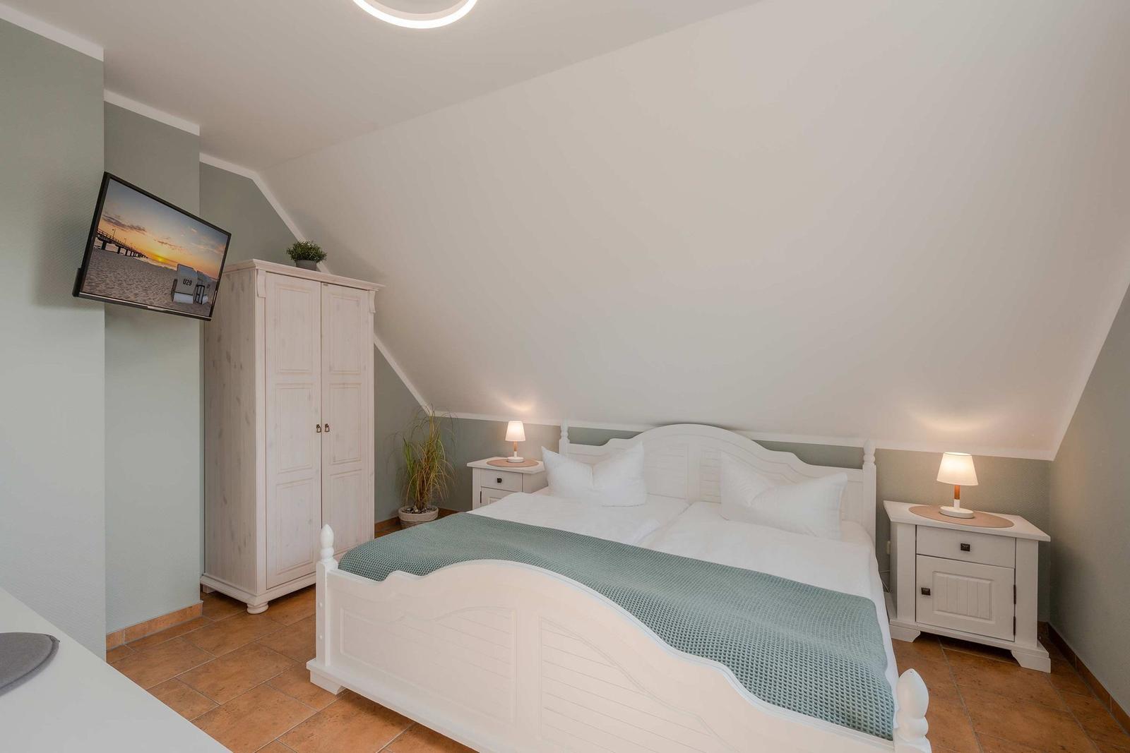 Schlafzimmer mit Dachschräge, weißem Bett, Nachttischen und TV an der Wand.