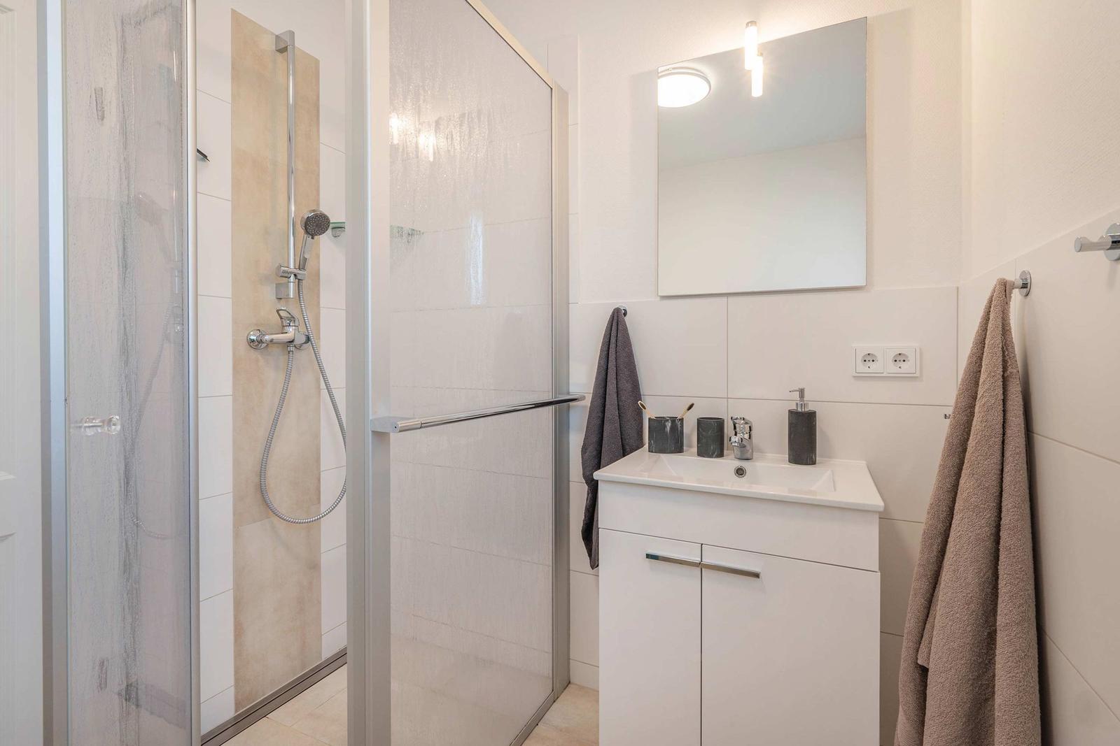 Badezimmer mit Dusche, Waschbecken und Spiegel. Handtuchhalter und Toilettenpapierhalter sichtbar.