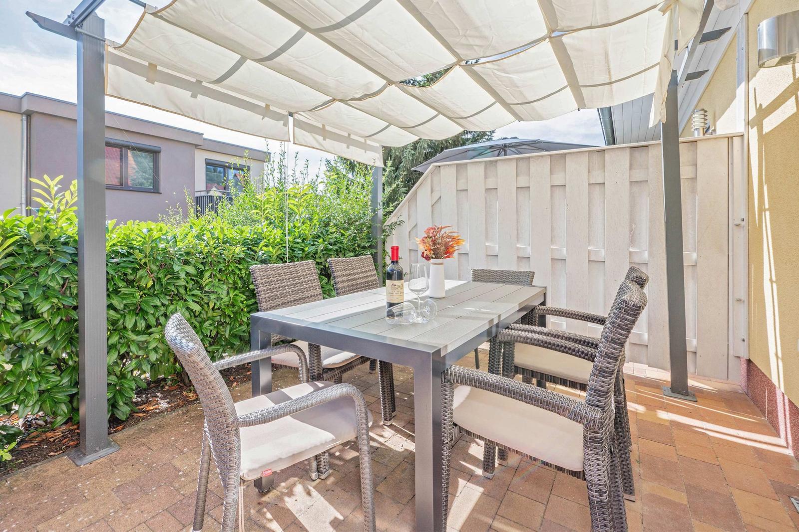 Terrasse mit Markise, Tisch und Stühlen. Auf dem Tisch steht eine Weinflasche und Gläser.