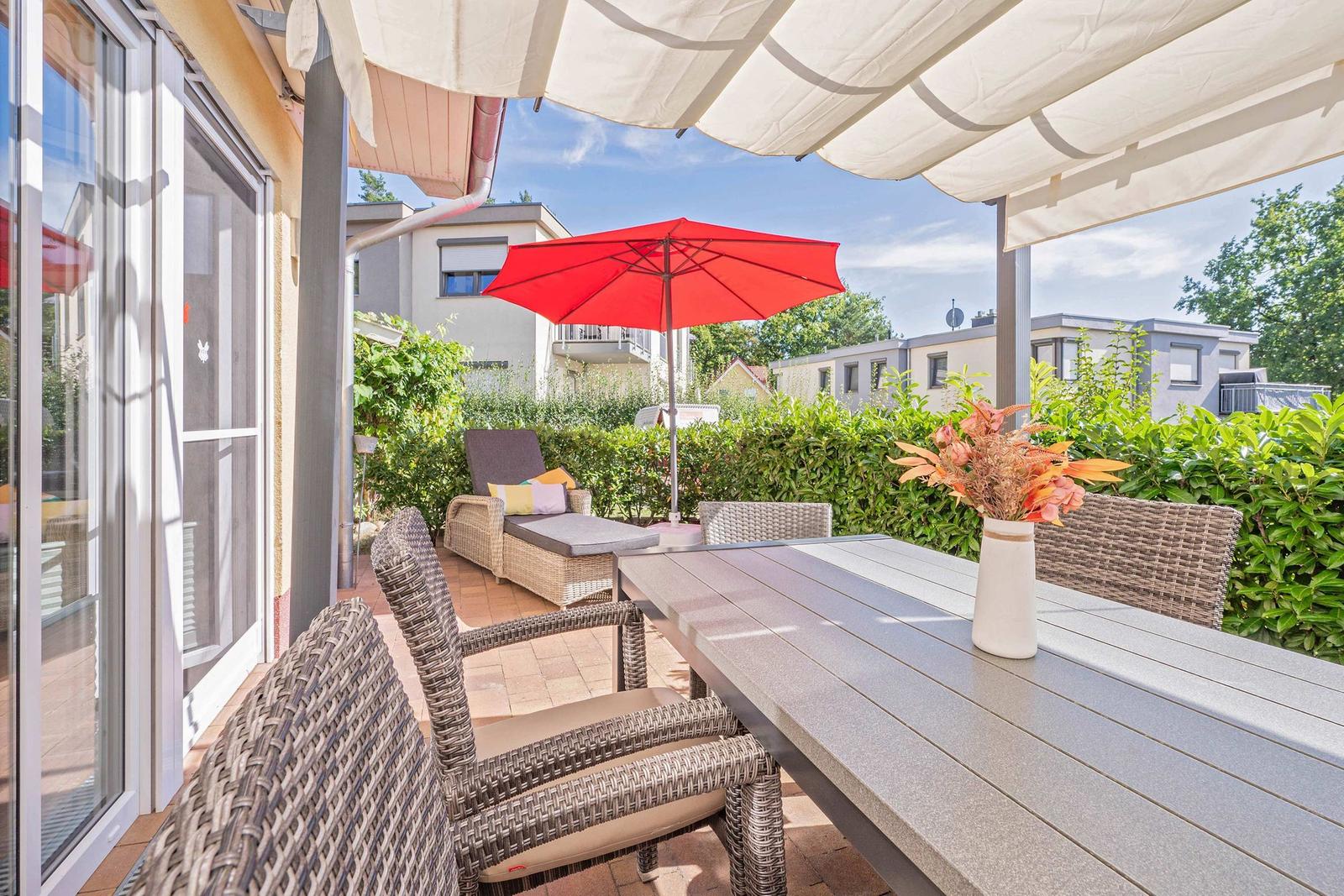 Terrasse mit Tisch, Stühlen und rotem Sonnenschirm. Umgeben von grüner Hecke.
