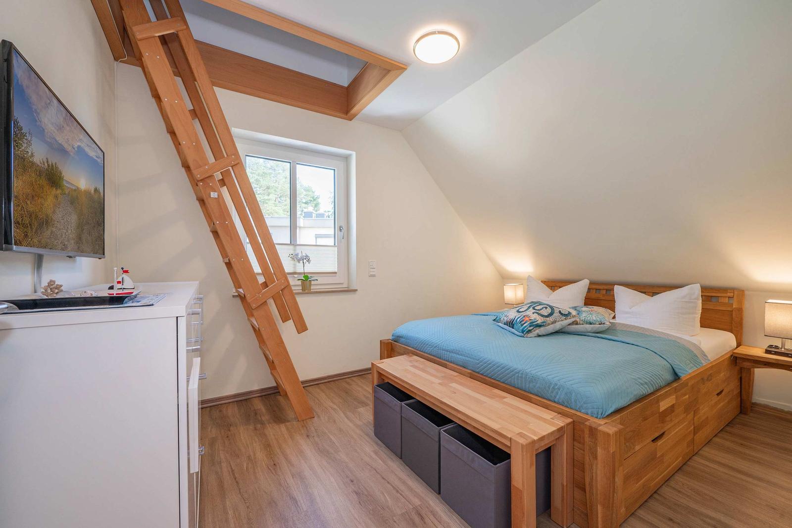 Schlafzimmer mit Dachschrägen, Holzbett, Fenster und Leiter zu Dachboden.