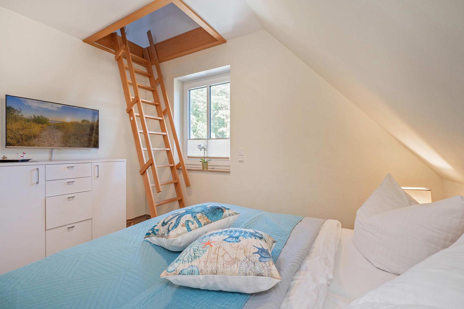 Schlafzimmer mit Dachschrägen, Holztreppe, Fenster und Bett mit Kissen.