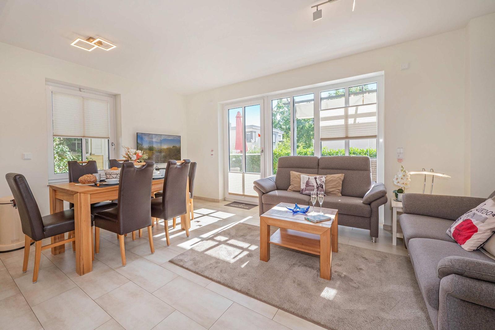 Wohn- und Essbereich mit Sofa, Tisch und Stühlen. Große Fenster mit Blick auf Balkon.