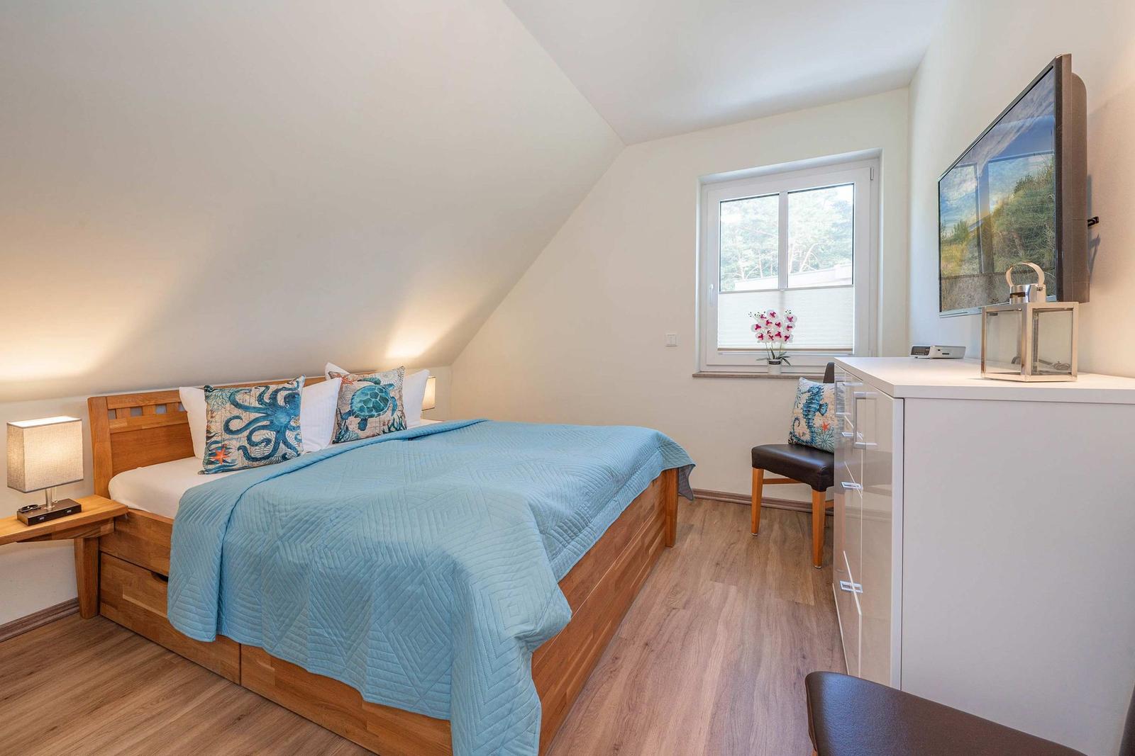 Schlafzimmer mit Dachschrägen, Bett mit blauer Decke und Fenster mit Blumen.