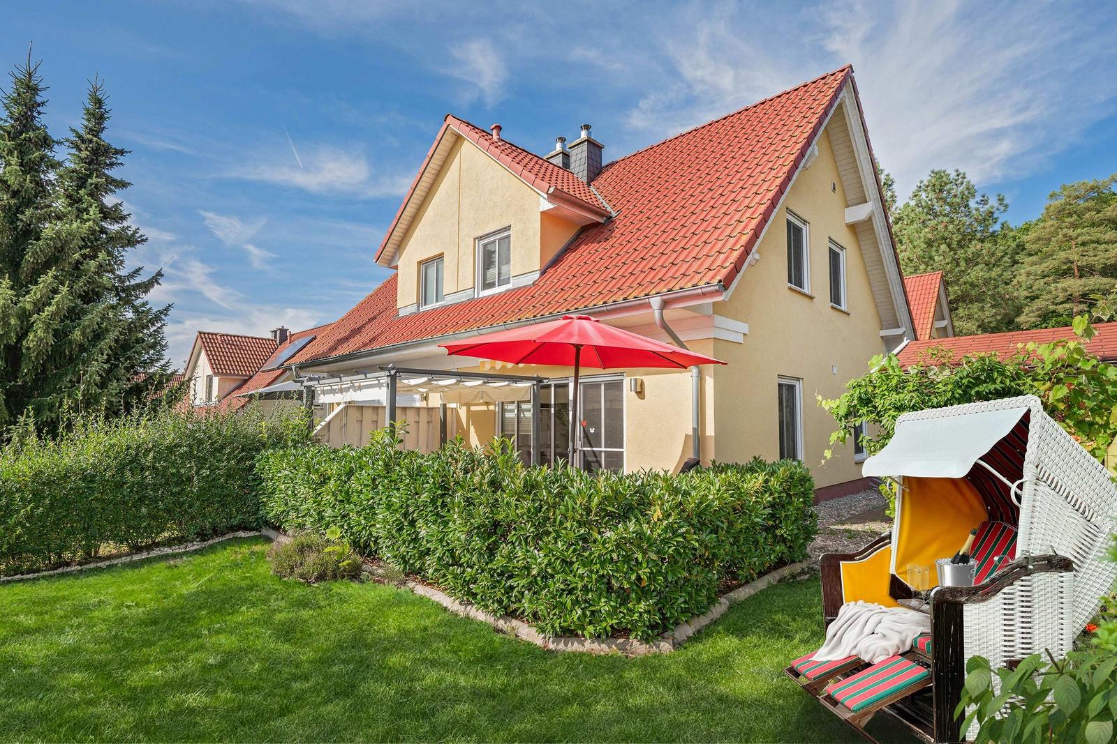 Haus mit Garten, Sonnenschirm und Liegestuhl. Roter Dachfirst, grüner Rasen.