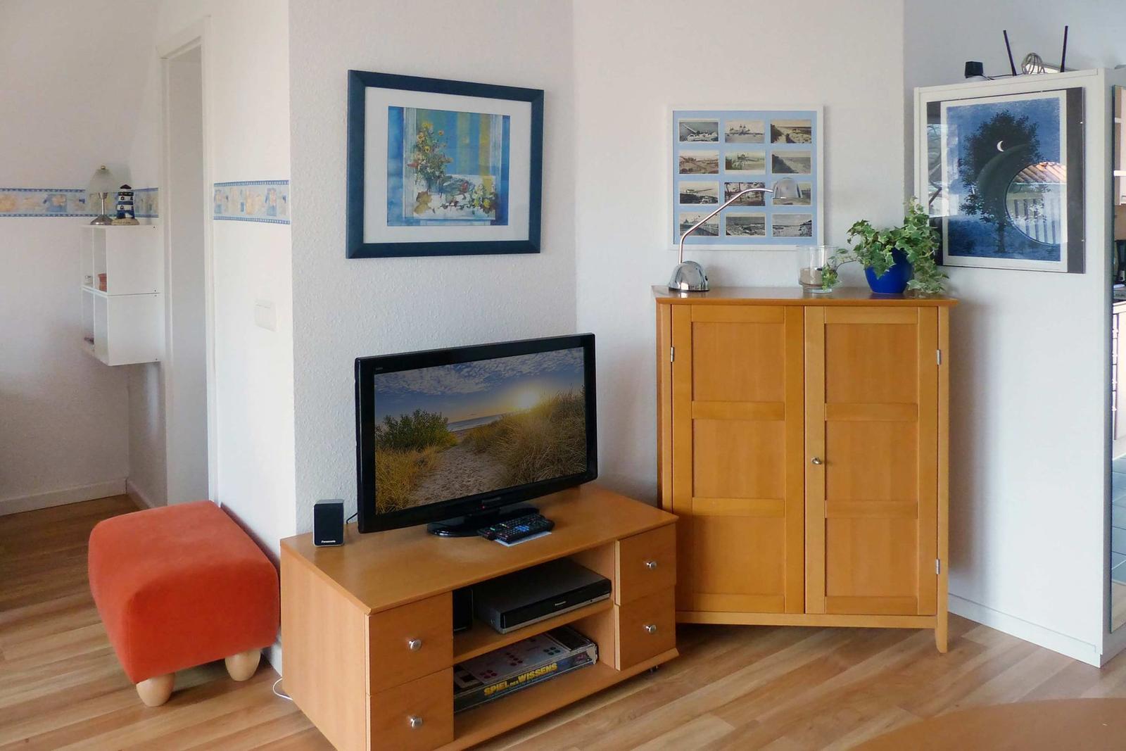 TV auf Holzregal, daneben Schrank und Wandbilder. Orange Sitzkissen links.