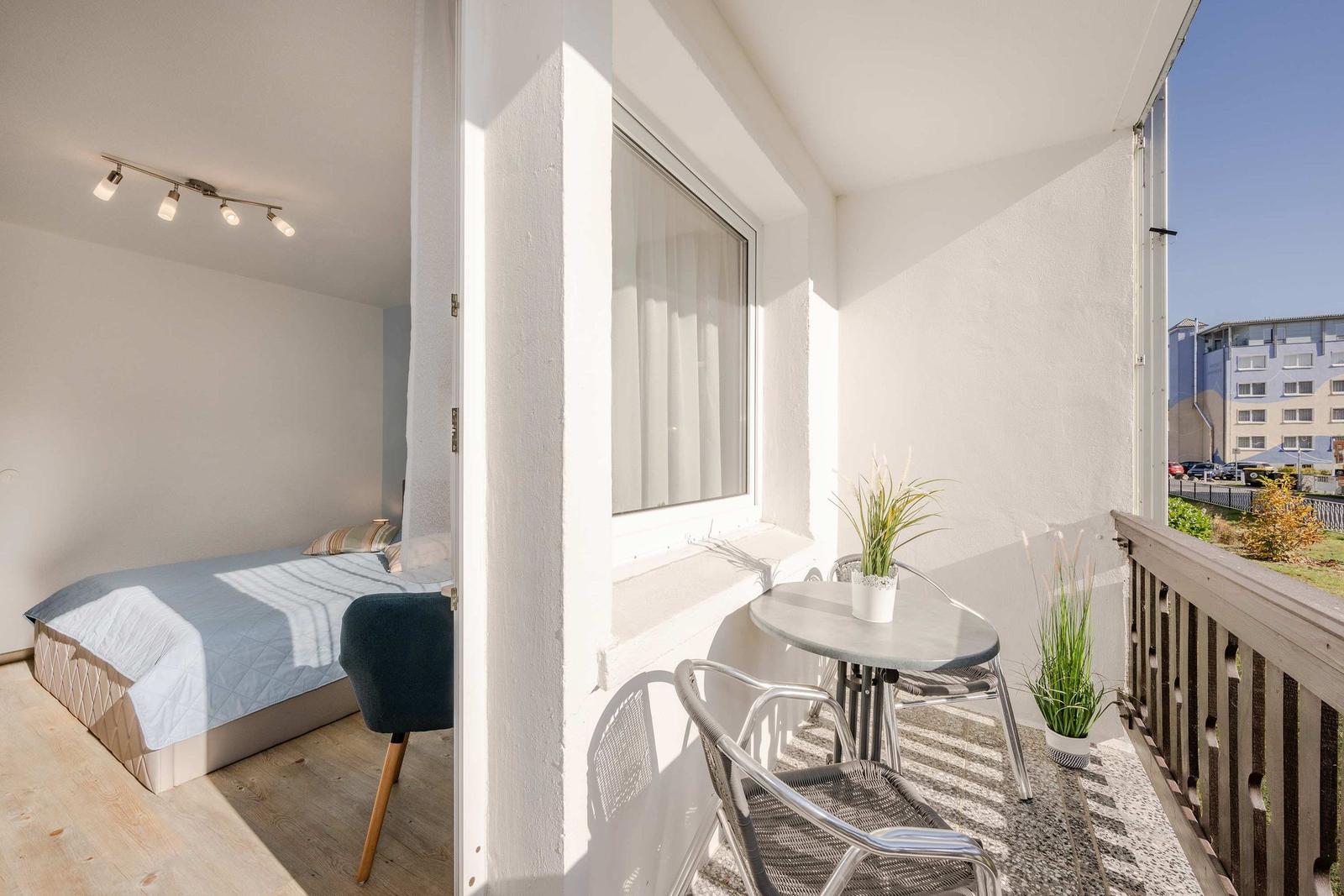 Ein Balkon mit Tisch und Stühlen. Im Hintergrund ist ein Schlafzimmer mit Bett und Stuhl zu sehen.