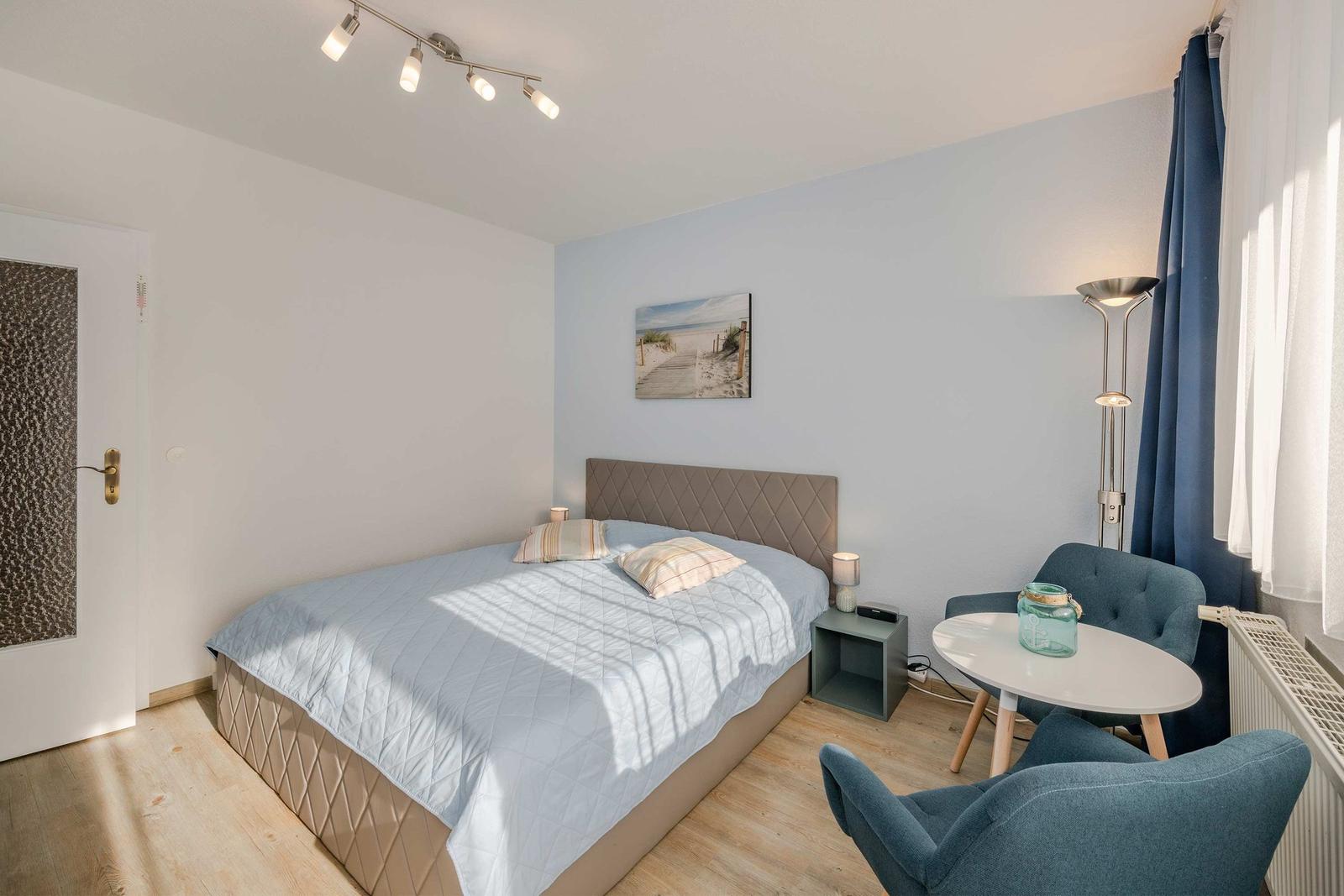Schlafzimmer mit Bett, zwei Stühlen und Tisch. Wandbild und Deckenleuchte sichtbar.