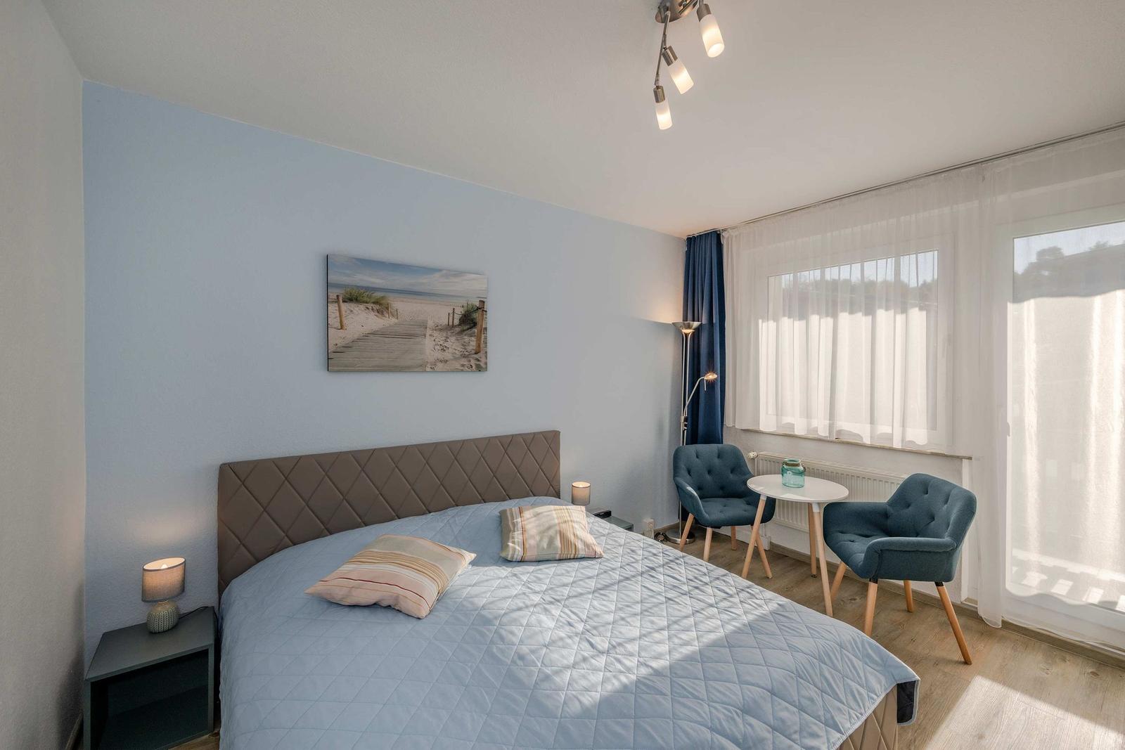 Schlafzimmer mit Bett, zwei Stühlen und Tisch. Wandbild zeigt Strand. Fenster mit Vorhängen.
