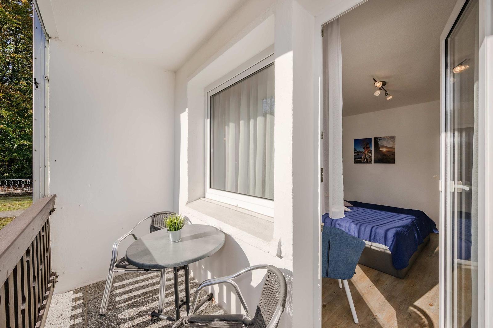 Balkon mit Tisch und Stühlen. Im Hintergrund Schlafzimmer mit Bett und Wandbildern.