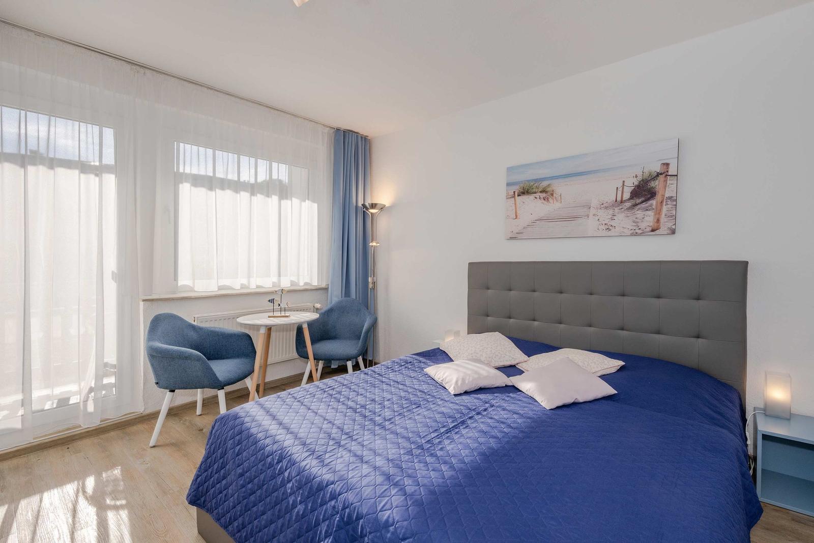 Schlafzimmer mit blauem Bett, zwei Stühlen und Fenster mit weißen Vorhängen.
