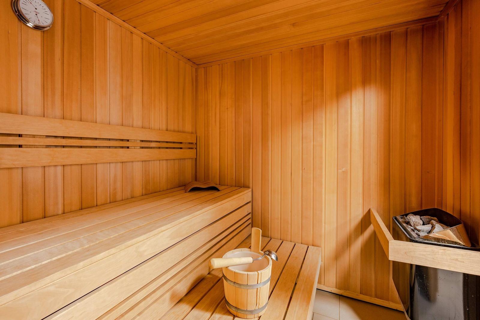 Sauna mit Holzbank, Eimer und Holzhammer, Wanduhr und Kachelofen sichtbar.