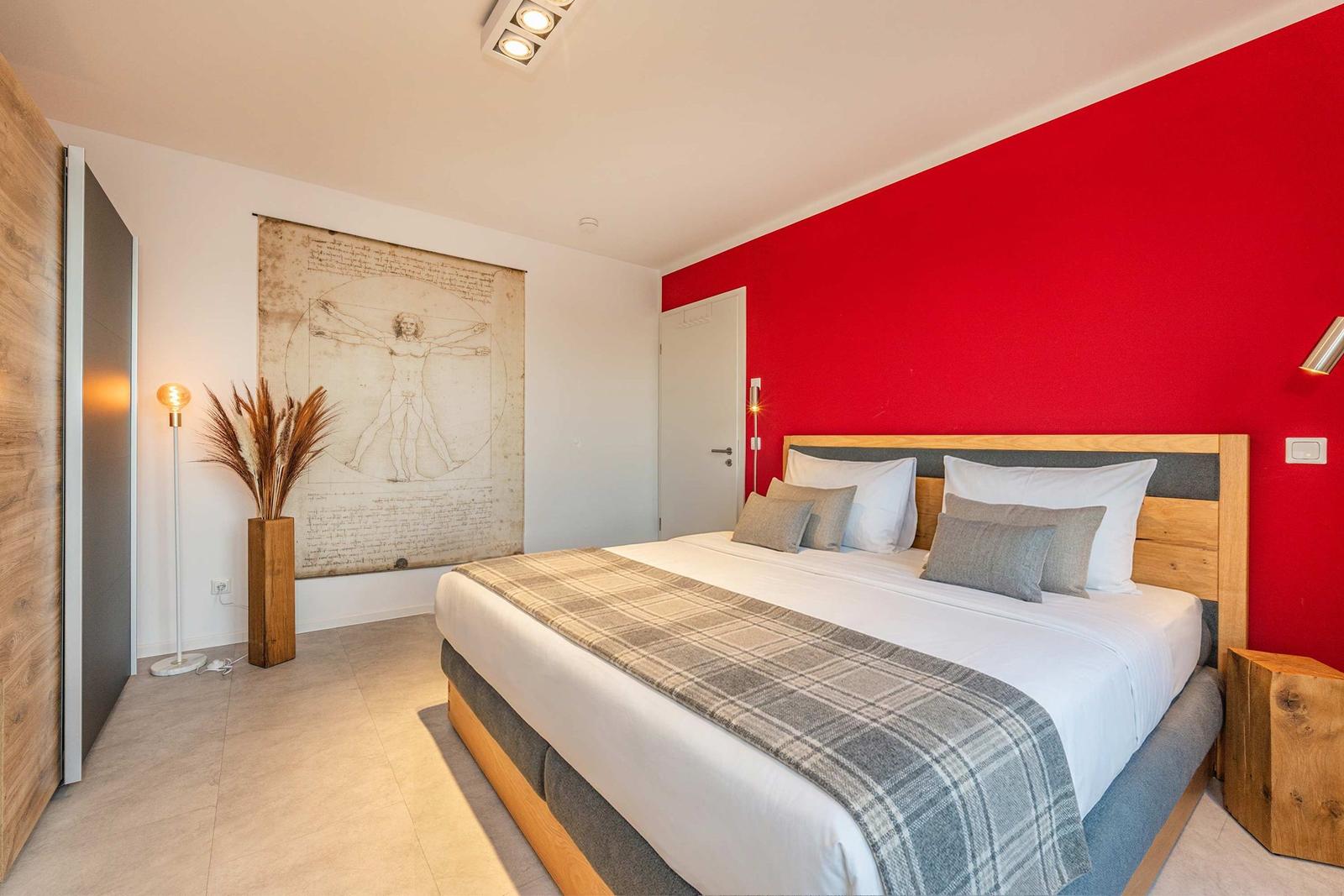 Schlafzimmer mit roter Wand, Bett mit Kissen und Decke, Wandbild und Stehlampe.
