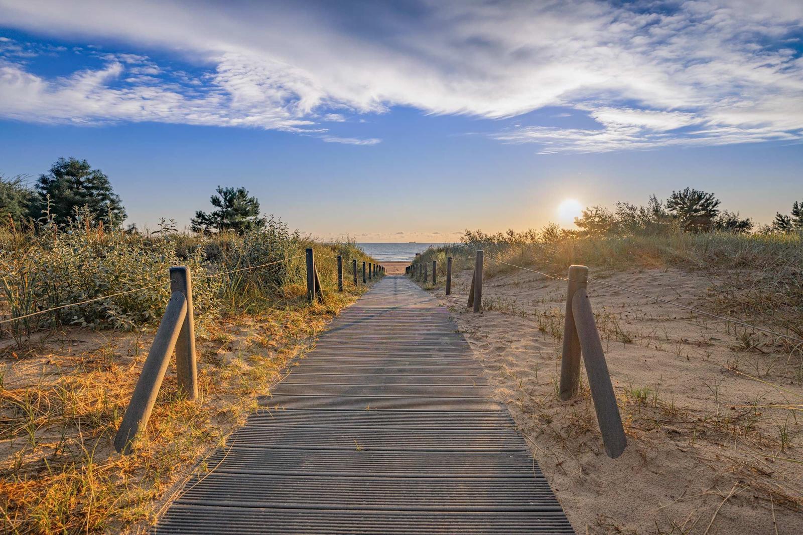 Holzweg führt durch Dünen zum Strand bei Sonnenuntergang.
