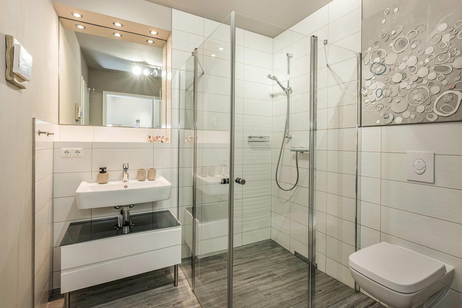 Badezimmer mit Dusche, Waschbecken und Toilette. Weiße Fliesen und Glasabtrennung.