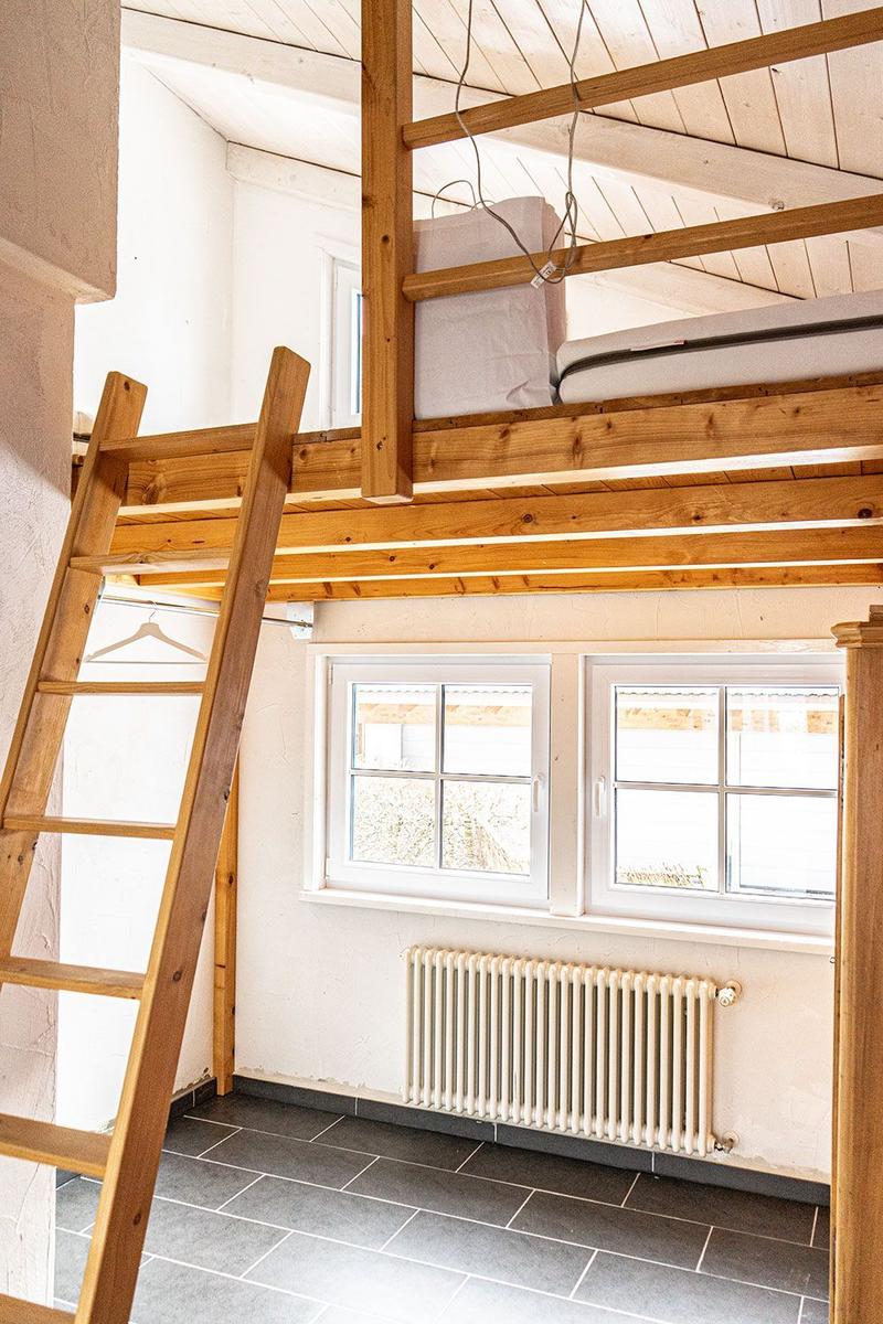 Upper bunk bed with wooden frame and ladder. Two windows and radiator on the wall.