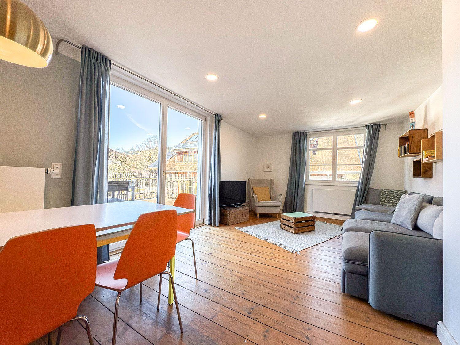 Living and dining area with wooden floor, sofa, TV, and view of balcony.