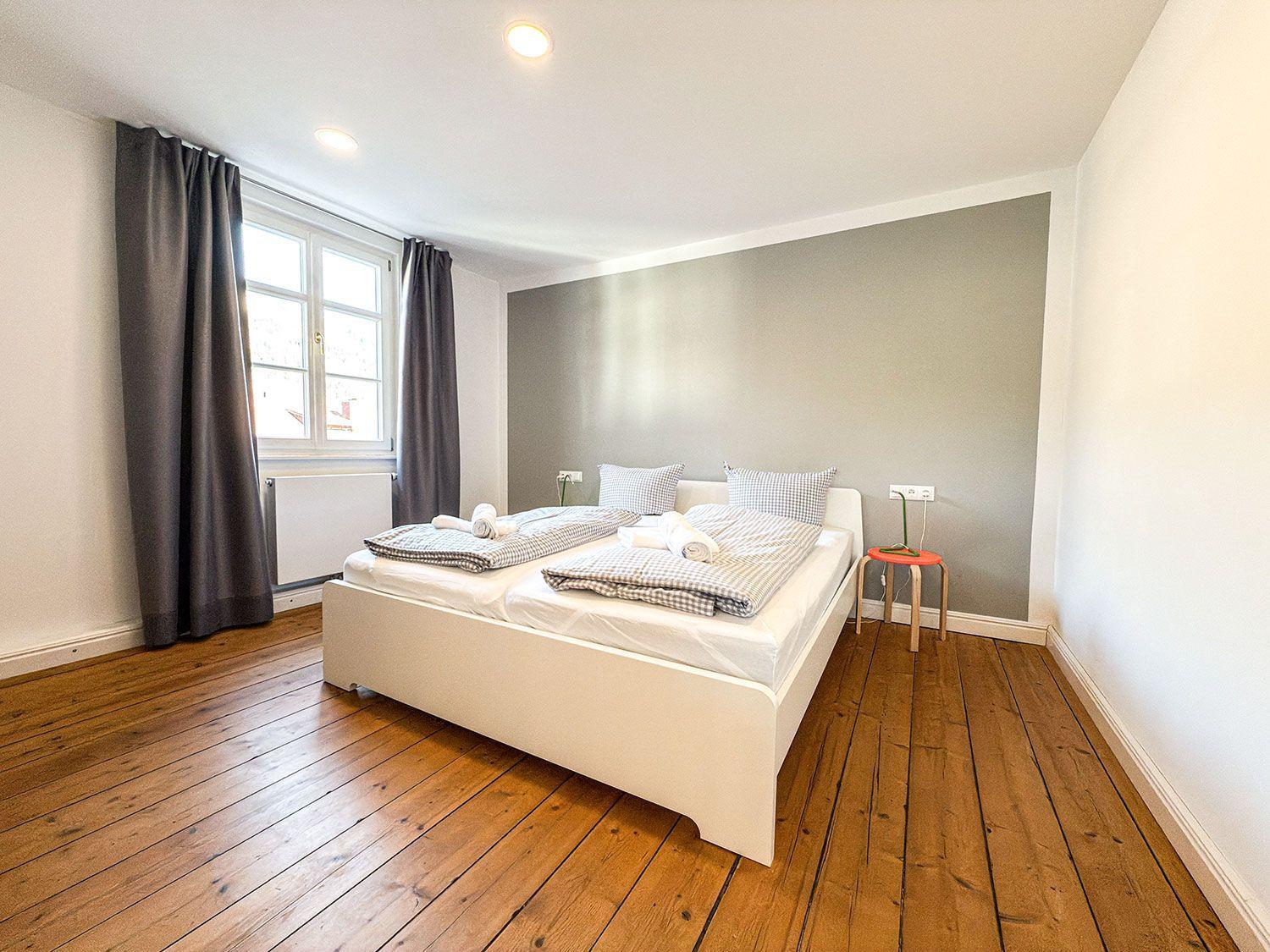 Bedroom with wooden floor, white bed, and gray wall. Window with dark curtains.