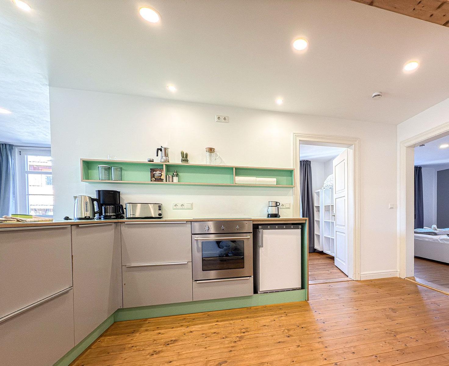 Kitchen with stainless steel stove, fridge, and wooden floor. View into bedroom and living area.