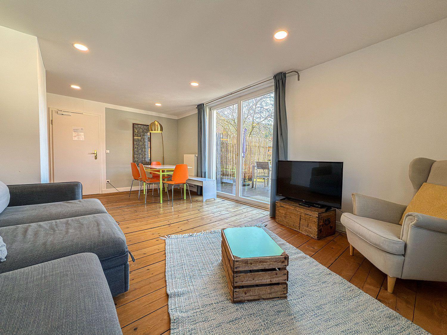 Living and dining area with wooden floor, sofa, armchair, and TV. Access to terrace with garden view.