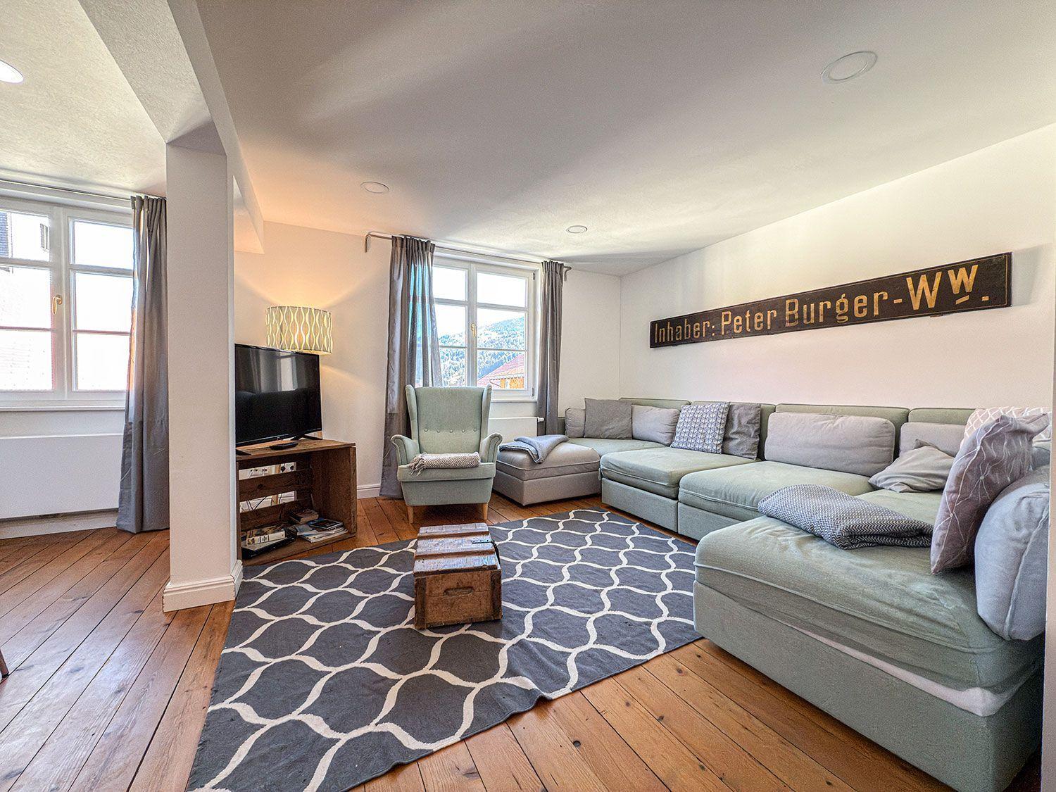 Wohnzimmer mit Sofa, Sessel und TV. Holzboden und großes Fenster.