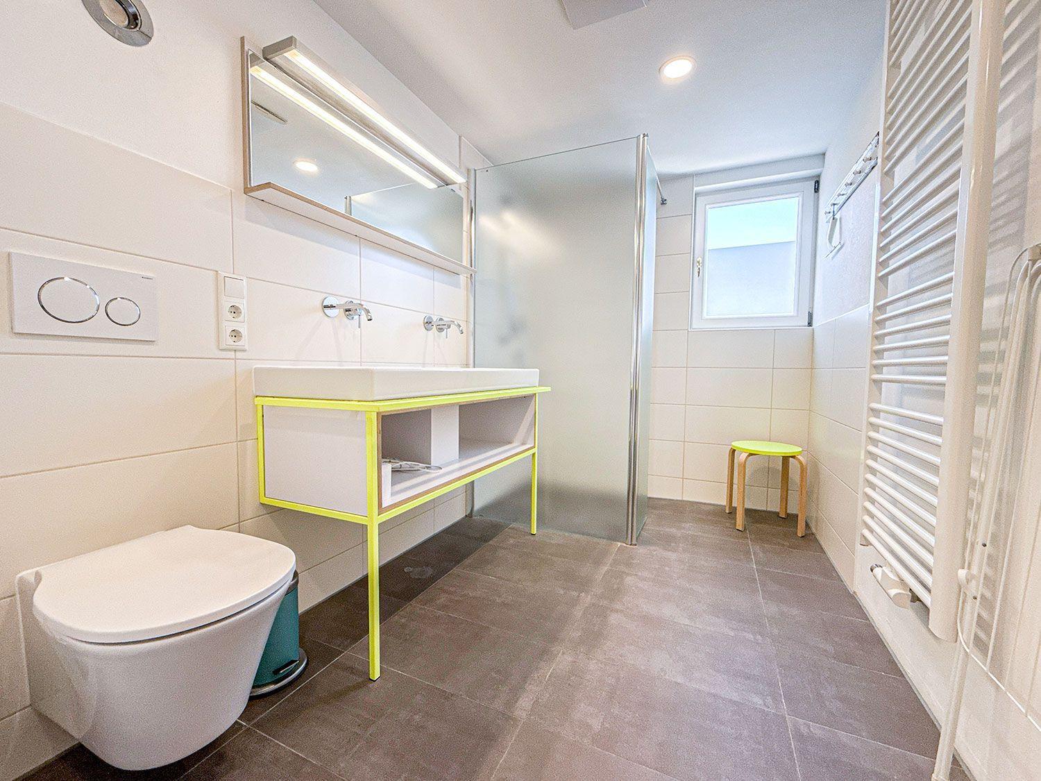 Badezimmer mit Dusche, Waschbecken und Toilette. Gelber Stuhl und Heizung.