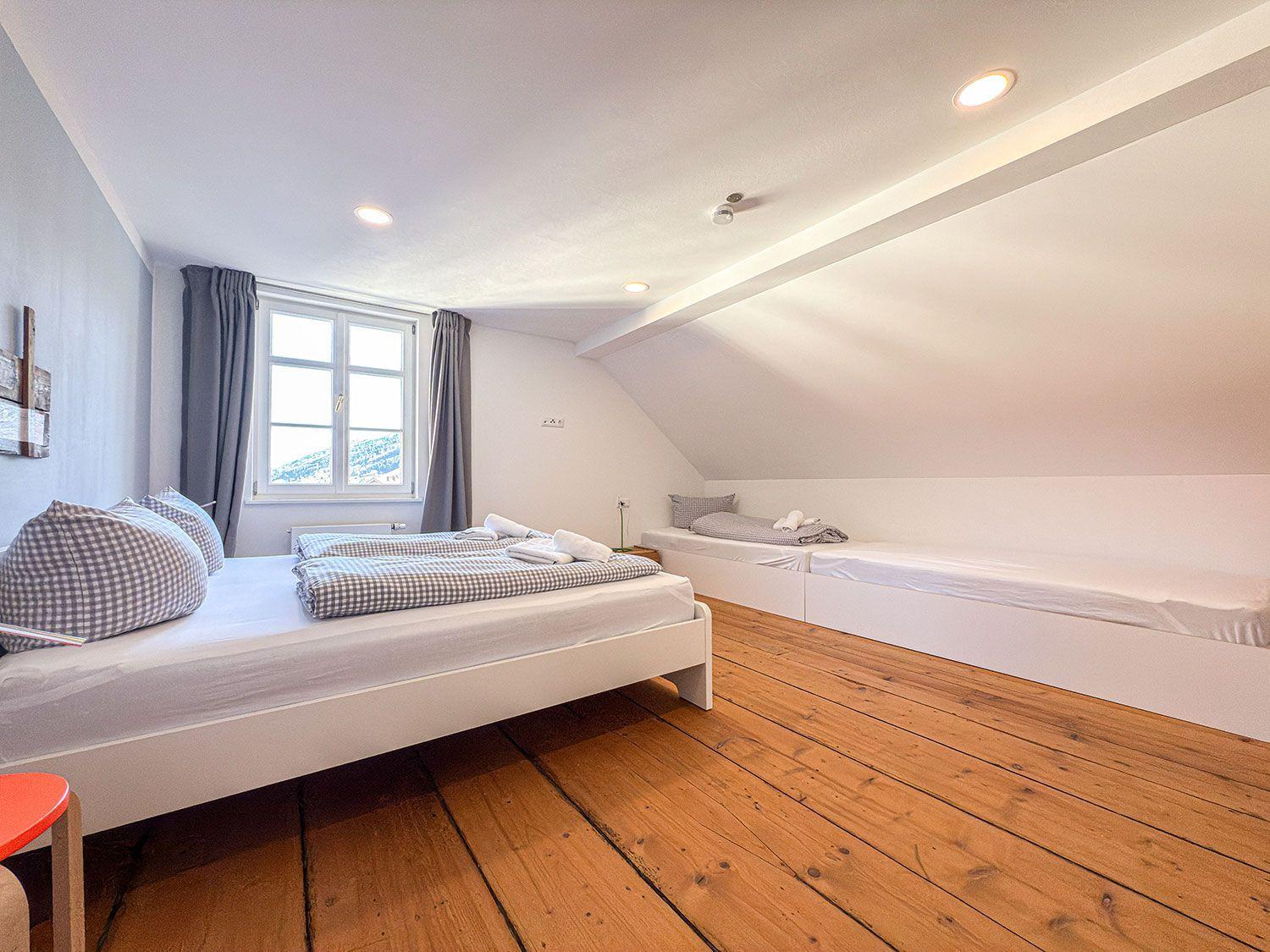 Zimmer mit Dachschräge, Holzboden und zwei Betten. Fenster mit Blick auf Berge.