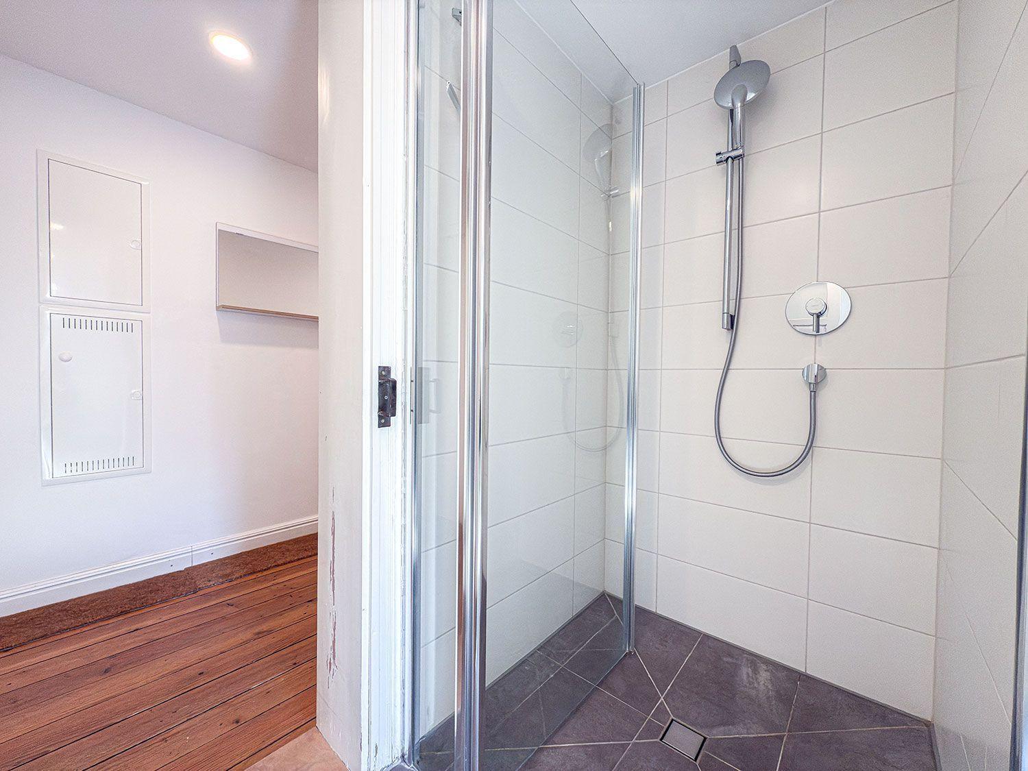 Dusche mit Glaswand, weißen Fliesen und Handbrause. Holzboden im angrenzenden Raum.