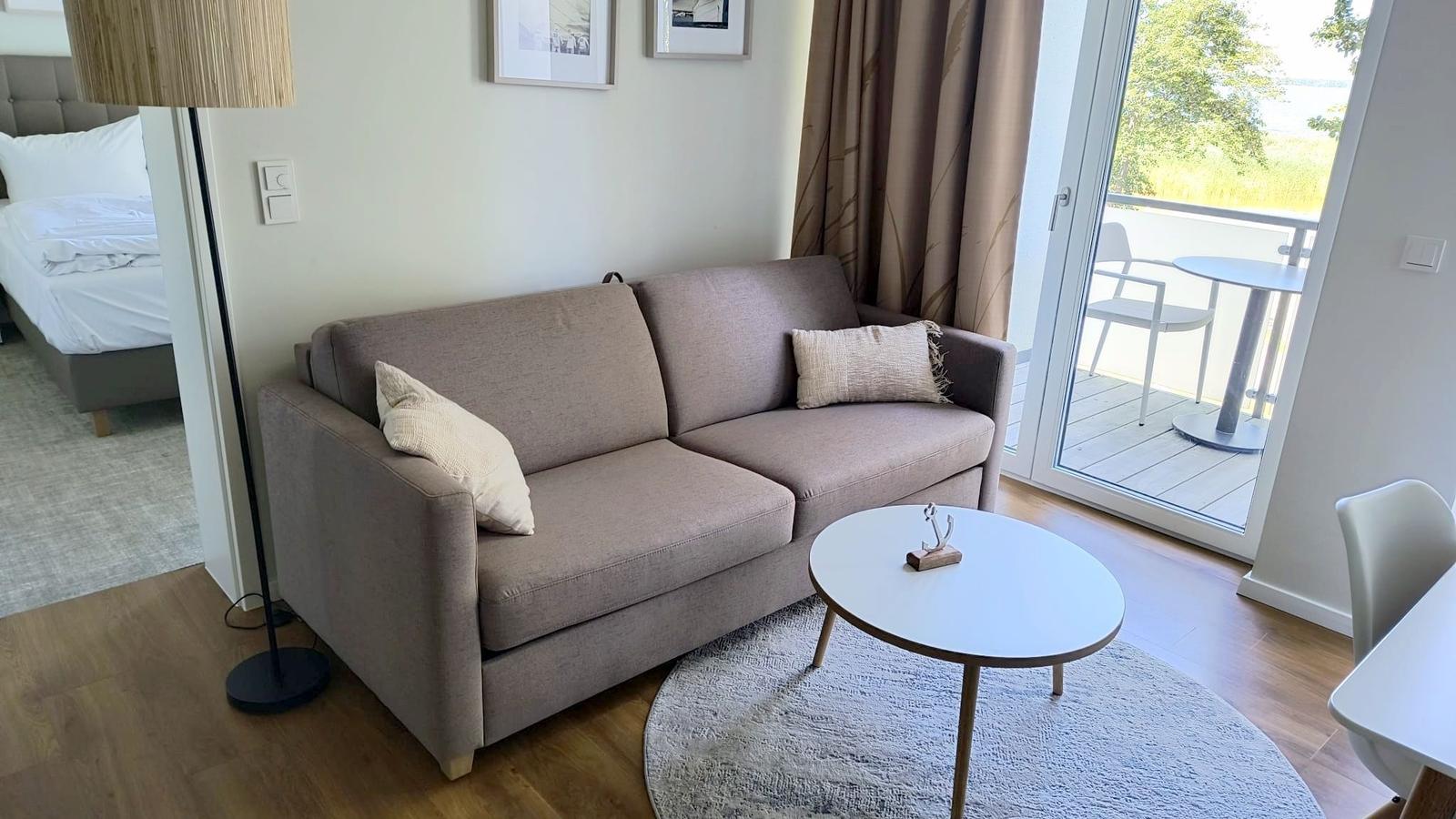 Ein grauer Sofa mit Kissen steht vor einer Glastür. Draußen ist eine Terrasse mit Tisch und Stühlen.
