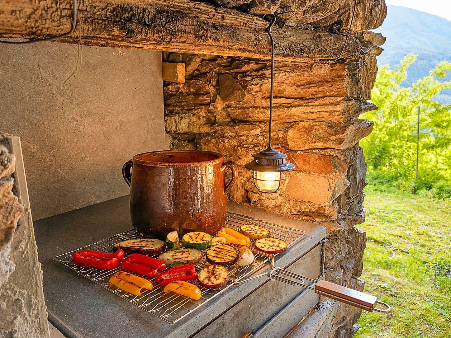 Grill with vegetables and pot against stone wall. Hanging lamp and view of the landscape.