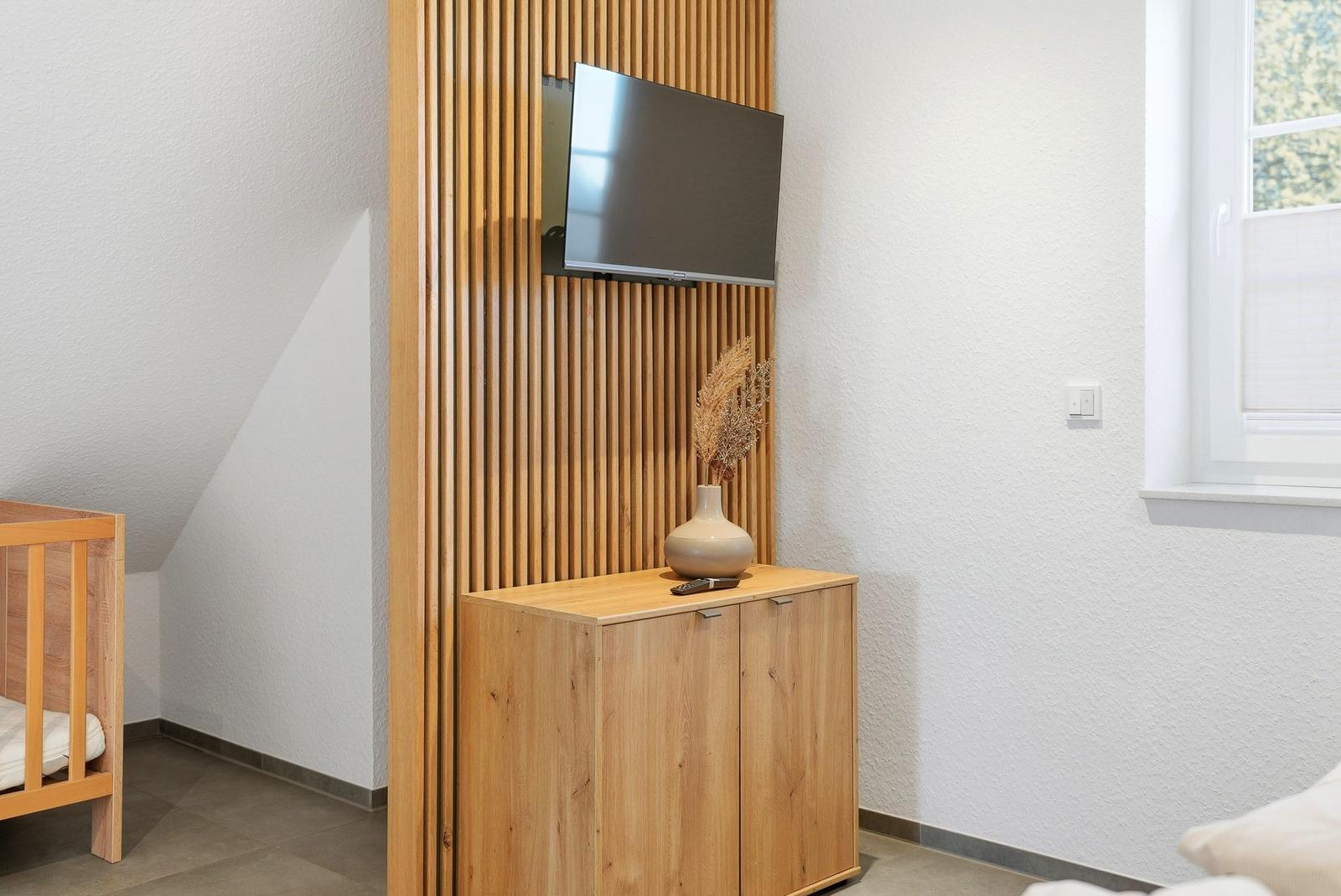 Schlafzimmer mit Holzschrank, Fernseher und Babybett. Fenster mit Jalousie.