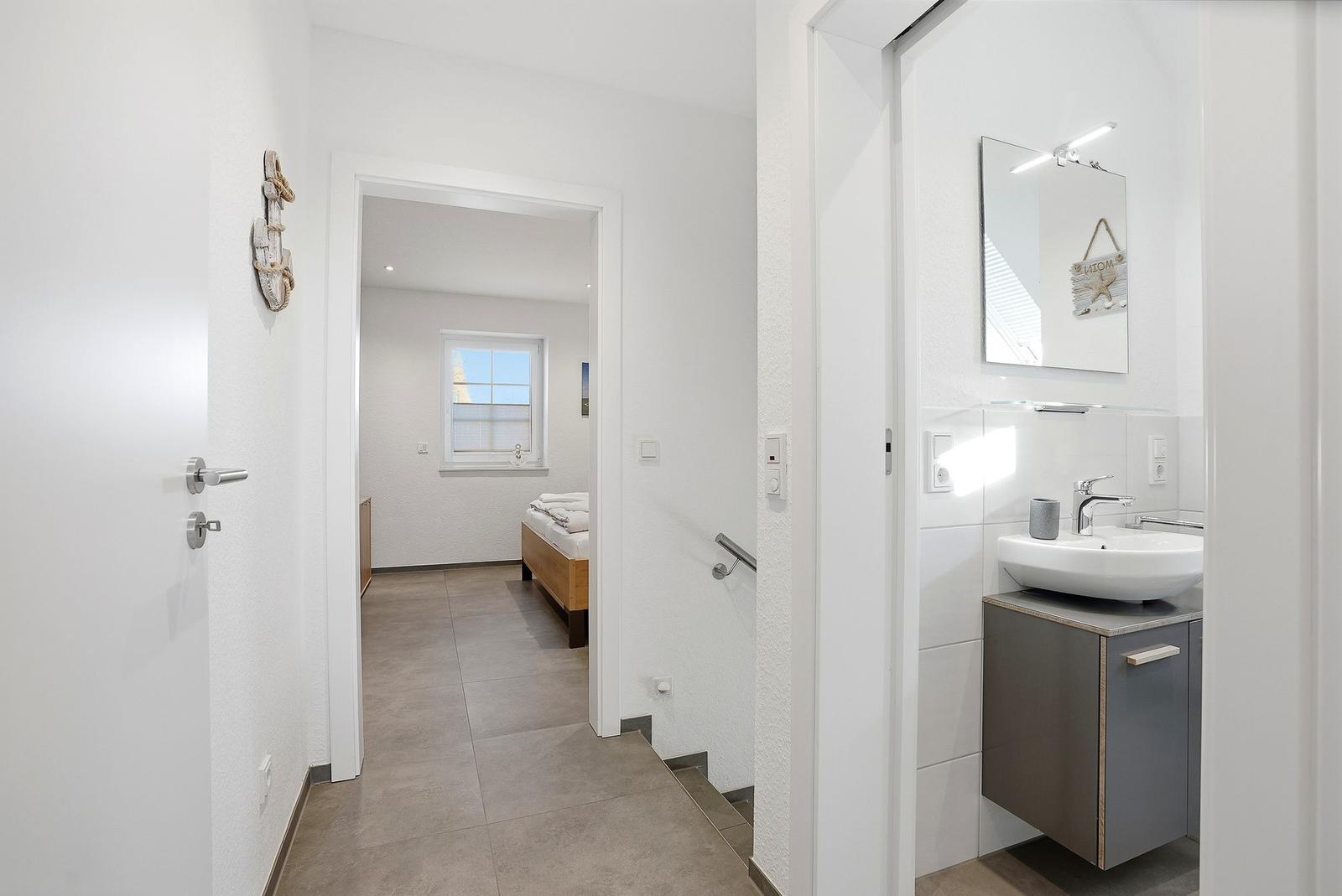Flur mit Bad und Schlafzimmer. Bad mit Waschbecken und Spiegel. Schlafzimmer mit Bett und Fenster.