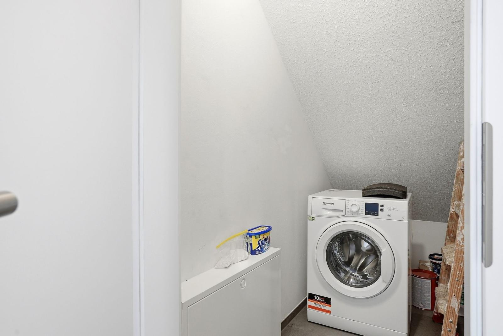 Waschmaschine in einem schrägen Raum mit weißen Wänden und einem kleinen Schrank.