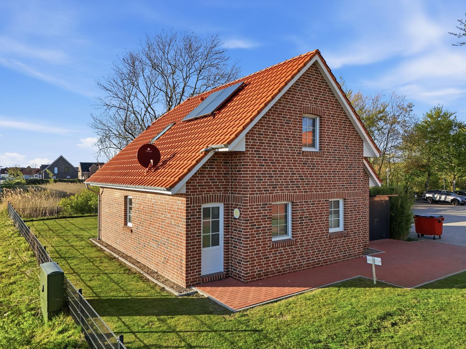 Ein zweistöckiges Haus mit rotem Ziegeldach und Backsteinfassade. Vor dem Haus befindet sich ein gepflasterter Bereich.