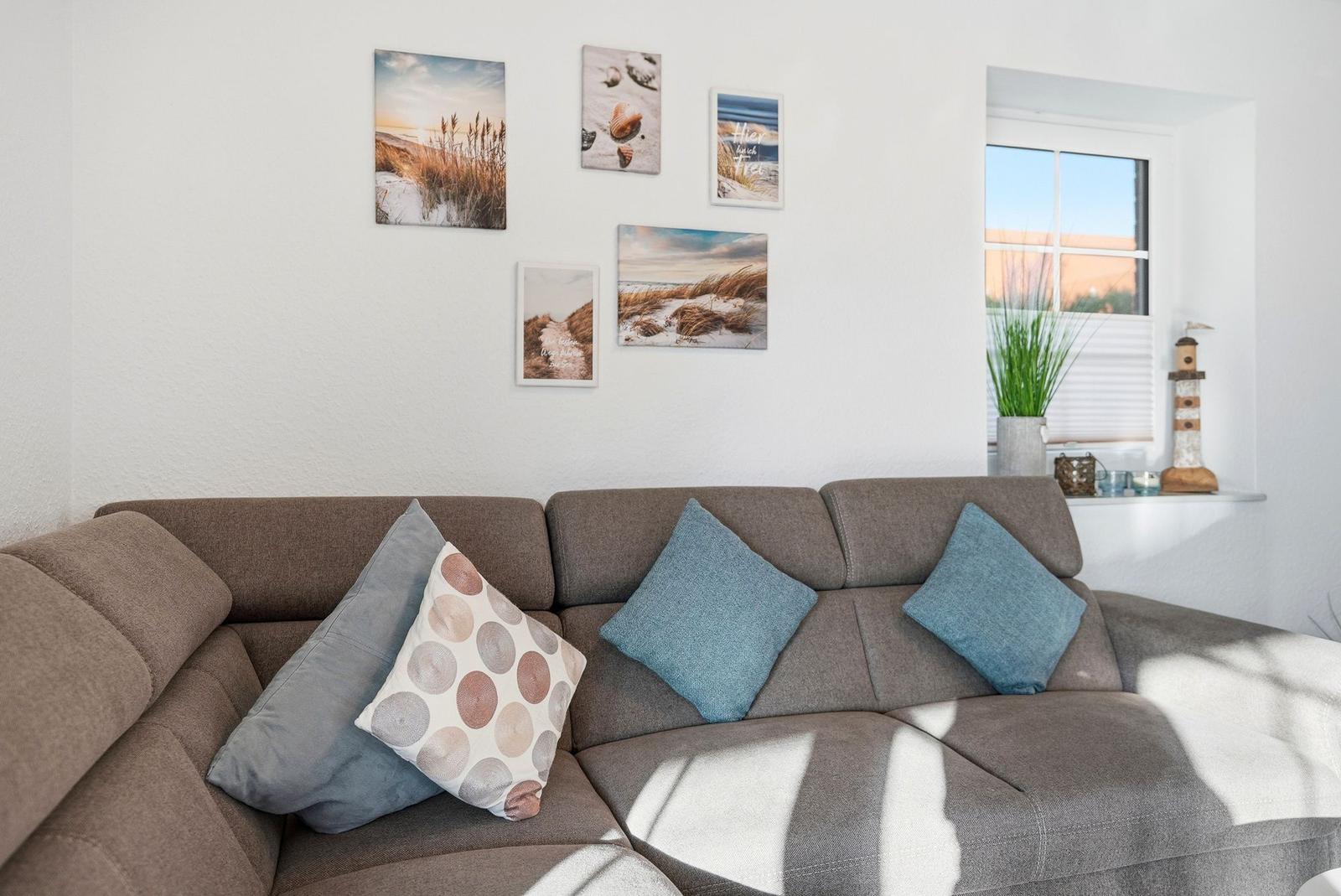 Ein grauer Sofa mit Kissen vor einer Wand mit Strandbildern und einem Fenster.