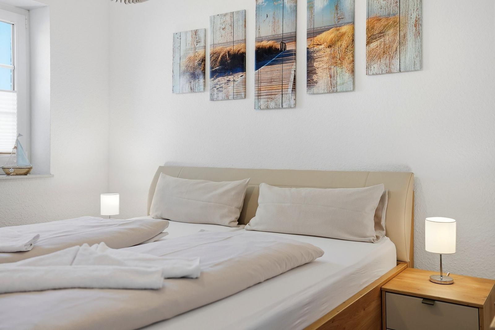 Schlafzimmer mit Bett, Nachttisch und Wandbildern. Fenster mit Segelboot-Dekoration.