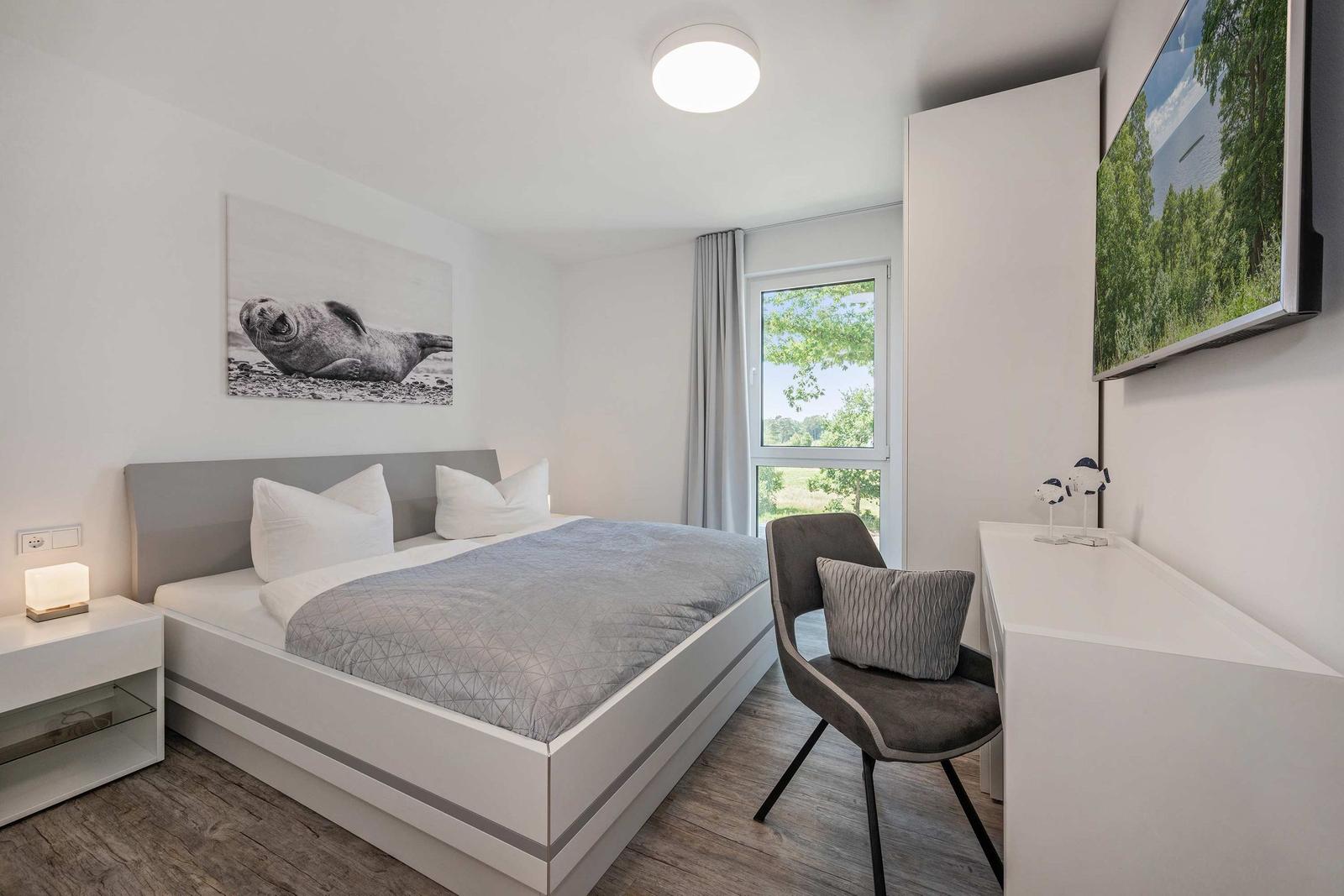 Schlafzimmer mit Doppelbett, Schreibtisch und Fensterblick auf Grünfläche.