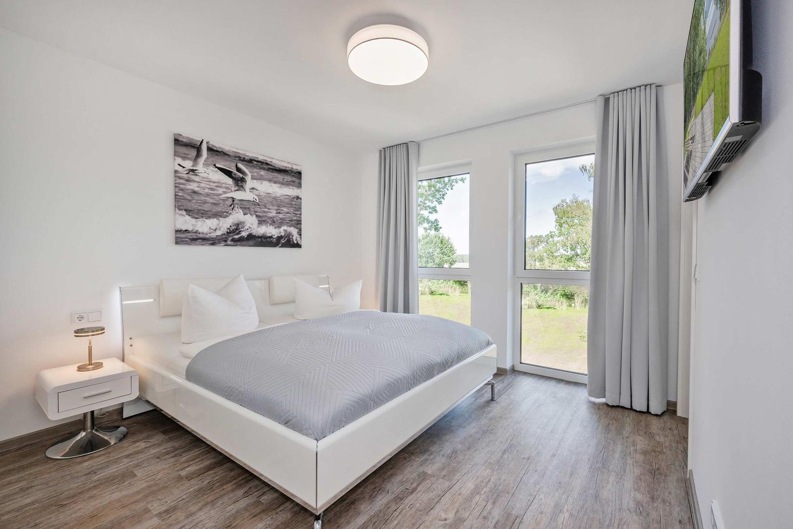 Schlafzimmer mit weißem Bett, Wandbild und Blick auf Grünflächen.