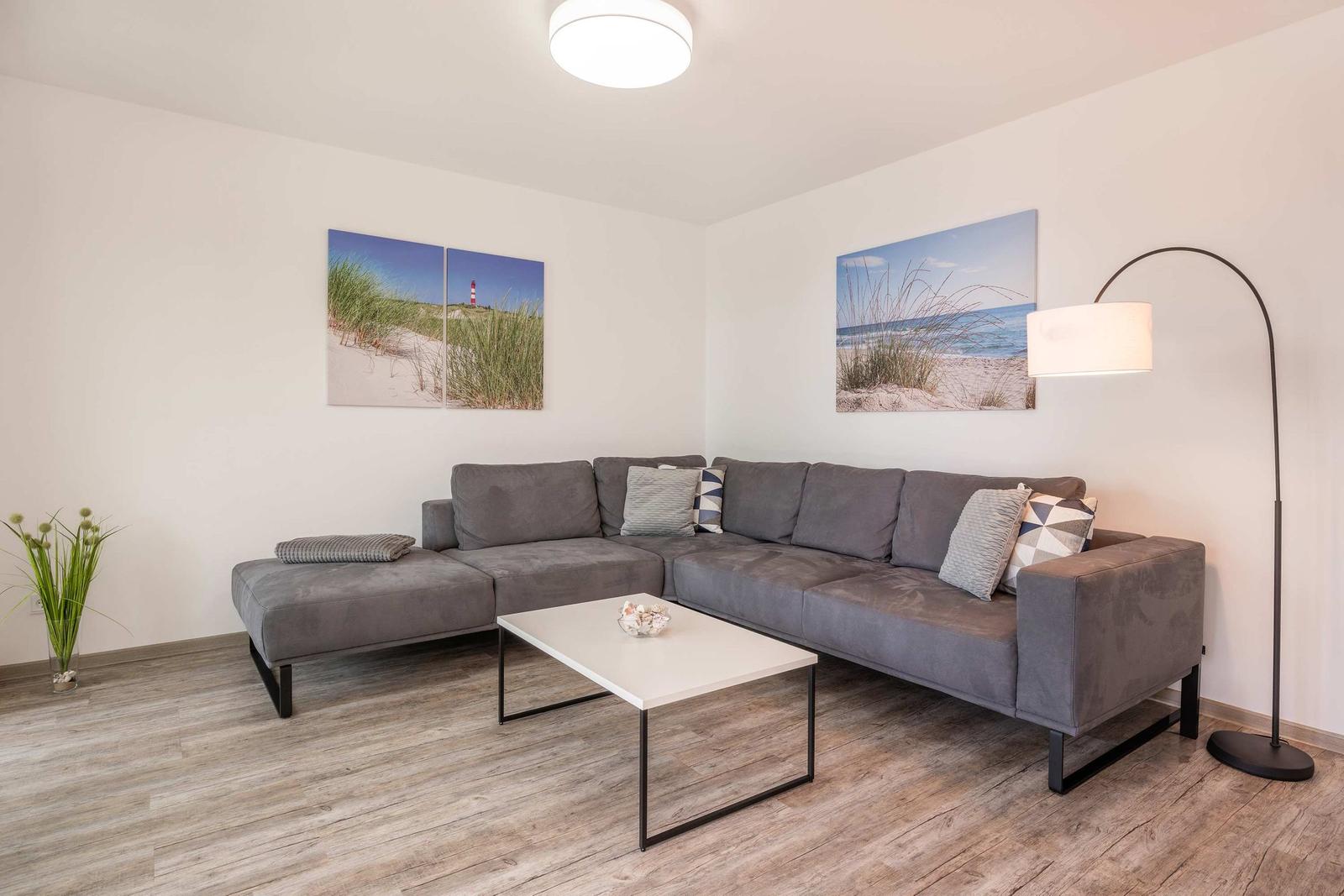 Ein Wohnzimmer mit grauem Sofa, weißen Wänden und Strandbildern an der Wand.