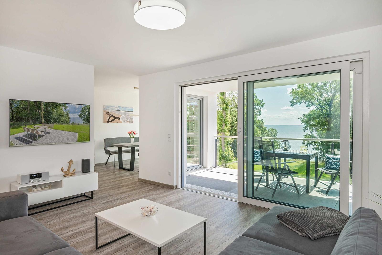 Wohnbereich mit Balkon, Fernseher und Essbereich. Blick auf Wasser und Bäume.