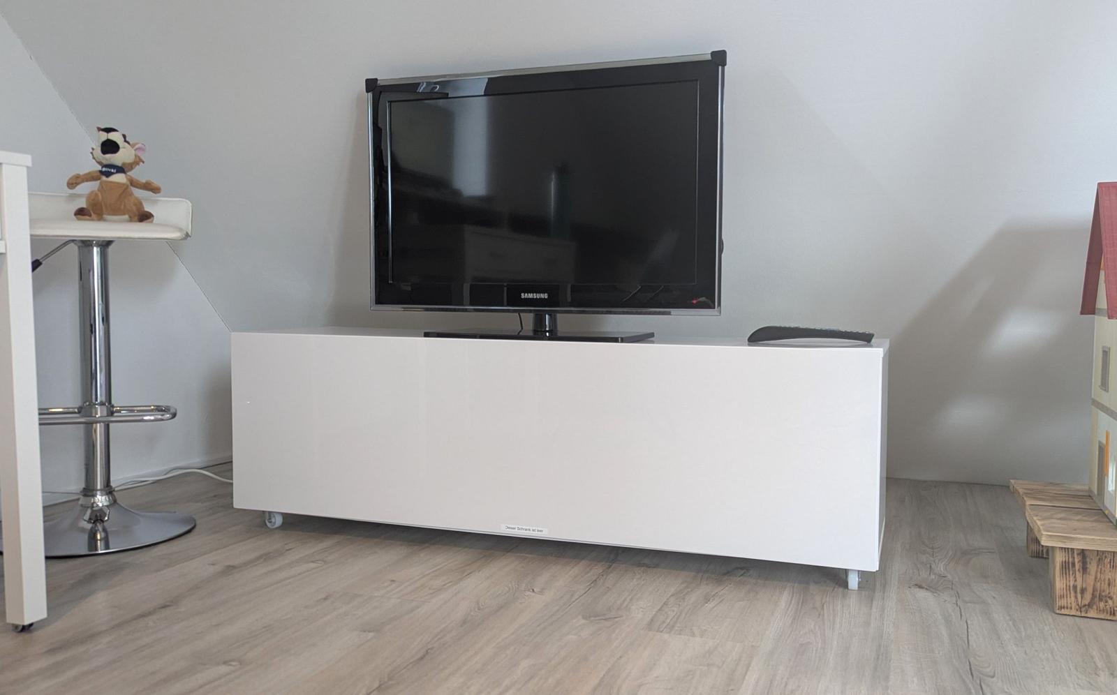 Ein Fernseher steht auf einem weißen TV-Schrank. Links ein Hocker mit Stofftier, rechts ein Holzhaus.