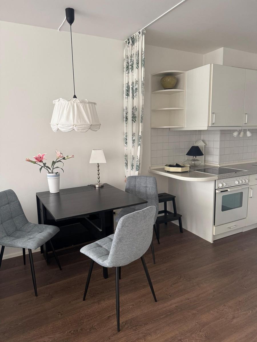 Dining area with table and chairs, kitchenette with stove and oven, light wall color and wooden floor.