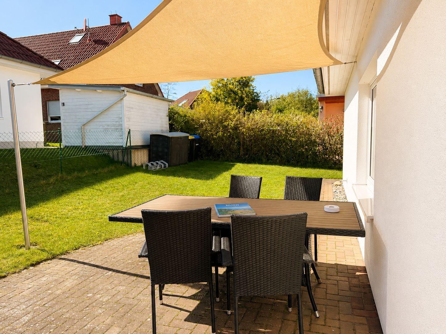 Terrasse mit Tisch und Stühlen unter Sonnenschutz, umgeben von Grünfläche und Hecke.