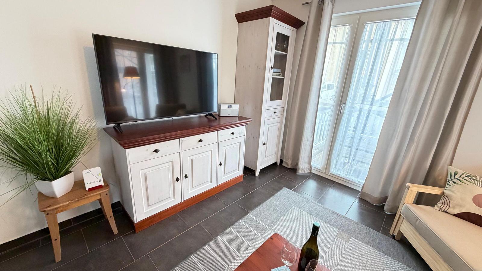 Wohnzimmer mit TV, Sideboard, Garderobe und Balkontür. Auf dem Tisch steht eine Weinflasche.
