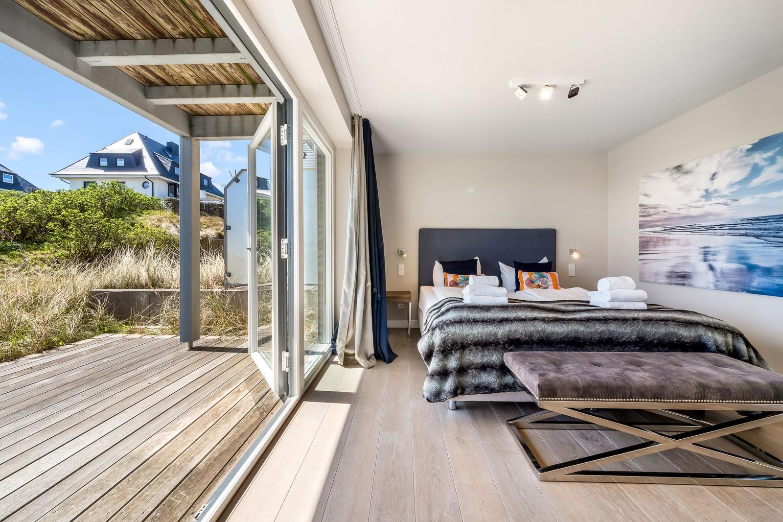 Schlafzimmer mit Blick auf Terrasse und Grünflächen. Bett mit Kissen und Decke. Wandbild und Sitzbank.